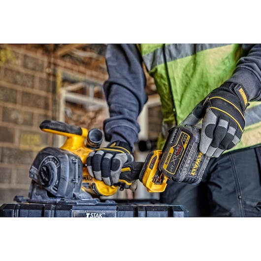 A person wearing protective gloves and a high-visibility vest is handling a DEWALT cordless power tool, specifically a planer, with a 18V XR FLEXVOLT battery, in a construction or workshop setting.