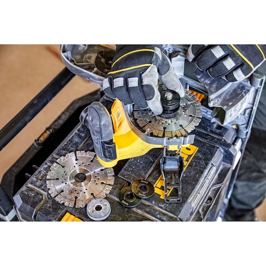Close-up of a person wearing protective gloves assembling or adjusting a circular cutting blade on a DEWALT power tool. Several metal blades and washers are arranged on a tool case.