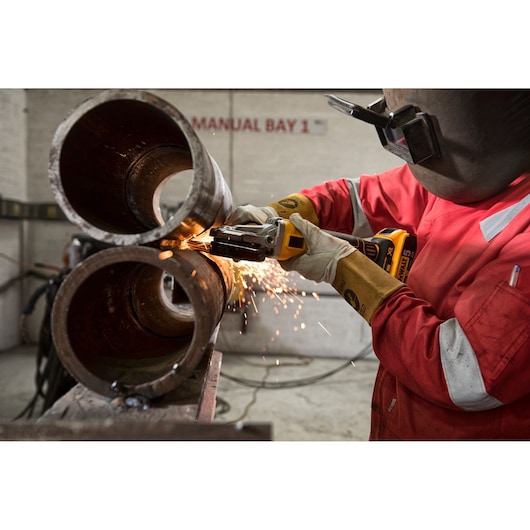 A worker wearing protective gear is using a DEWALT cordless angle grinder (model DCG405FN) to cut through large metal pipes indoors. Bright sparks are visible as the grinder operates.