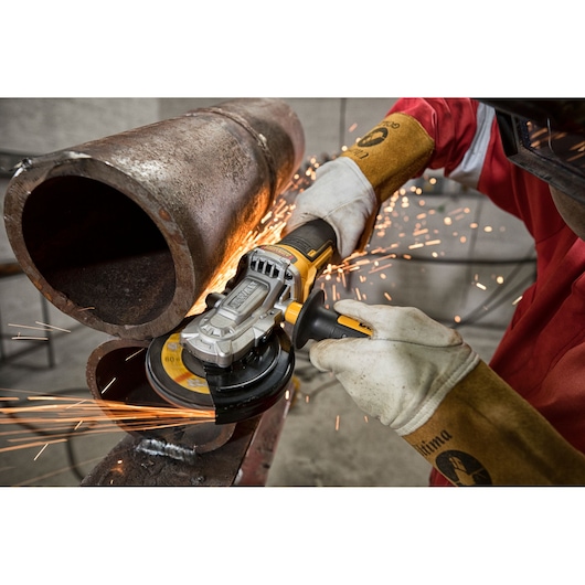 A person wearing protective gloves and clothing uses a DEWALT angle grinder (DCG405FN) to cut a large metal pipe, producing sparks in an industrial setting.