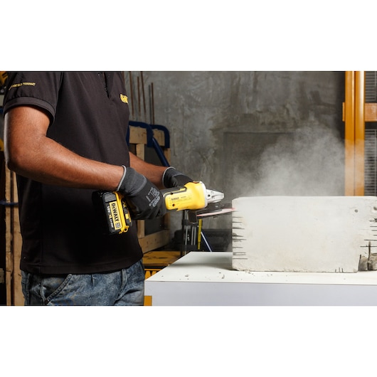 A person wearing gloves using a DEWALT cordless angle grinder to cut or grind concrete, with visible dust rising from the work area.