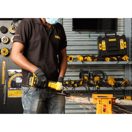 Person wearing a mask and gloves using a DEWALT cordless angle grinder to cut a metal rod, with sparks flying. Various DEWALT power tools and accessories are displayed in the background.