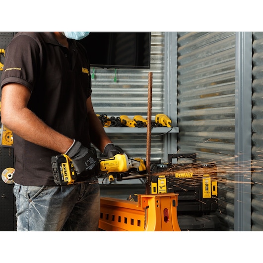 A person wearing a black DEWALT shirt and gloves uses a yellow DEWALT cordless angle grinder to cut a metal rod, with sparks flying. The scene is indoors, with other DEWALT tools visible in the background.