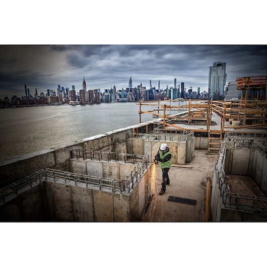 Worker uses power tool on concrete at construction site near waterfront, city skyline in background.