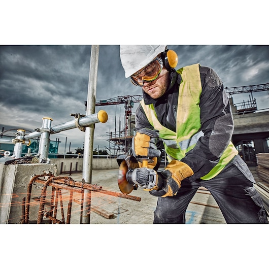 A construction worker wearing a safety vest, gloves, and a hard hat uses a DEWALT angle grinder to cut through rusty rebar at a construction site. The worker is focused on the task, with sparks flying from the cutting tool and scaffolding, structures, and cranes in the background.