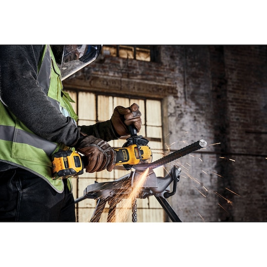 A worker wearing safety gear uses a DEWALT cordless grinder to cut through a metal rod, producing sparks, in an industrial setting.