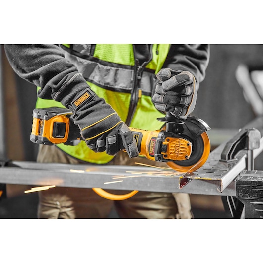 A person wearing gloves and a safety vest uses a DEWALT cordless angle grinder to cut through a metal piece, with sparks flying from the cutting area.