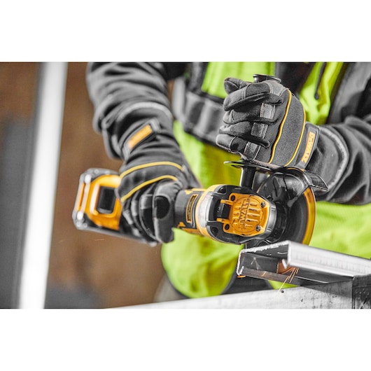 A person wearing protective gloves and a yellow safety vest is using a DEWALT cordless angle grinder (model DCG409NT) to cut a piece of metal.