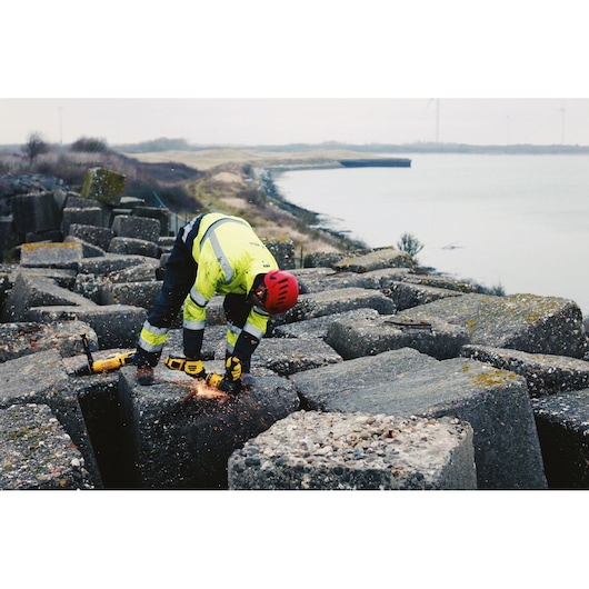 A person wearing a safety helmet and high-visibility jacket uses a yellow DEWALT DCG414N angle grinder to cut into a large concrete block outdoors near a body of water, with sparks visible during operation.