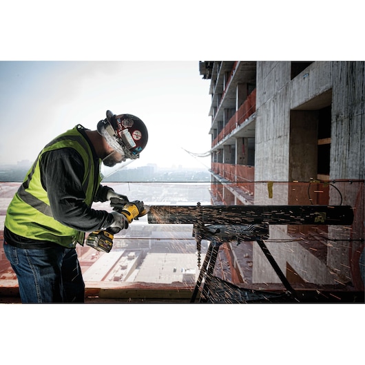 A construction worker wearing a safety vest, gloves, and a helmet is using a DEWALT cordless grinder to cut a metal pipe on a building site. Sparks are flying from the pipe, and a partially constructed concrete building is visible in the background.
