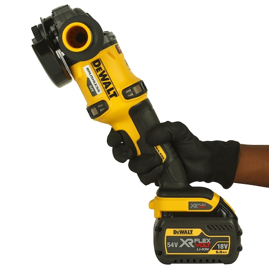 A person wearing a black glove is holding a DEWALT cordless angle grinder with a brushless motor and XR FLEXVOLT lithium-ion battery, model DCG414T2, against a white background.