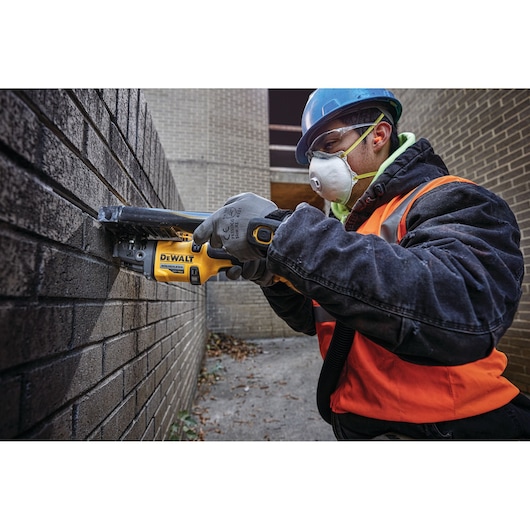 A person wearing a blue hard hat, orange safety vest, and gloves uses a DEWALT DCG418B power tool on a brick wall at an outdoor construction site. The face of the person is blurred.