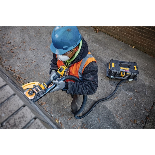 A worker wearing a blue hard hat and orange safety vest is using a DEWALT power tool attached to a dust extraction unit, grinding the edge of a concrete surface outdoors.