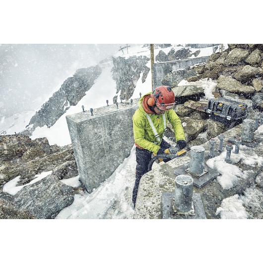 A worker wearing a helmet and safety gear uses a DEWALT power tool outdoors at a snowy construction site surrounded by rocks