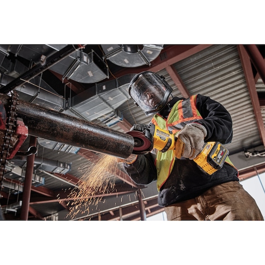 A worker wearing safety gear uses a DEWALT DCG418NT cordless angle grinder to cut a metal pipe, producing sparks in an