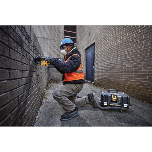A construction worker wearing a blue safety helmet, orange vest, and gloves is kneeling outdoors using a DEWALT power tool on a brick wall. A DEWALT tool case is placed on the ground nearby.