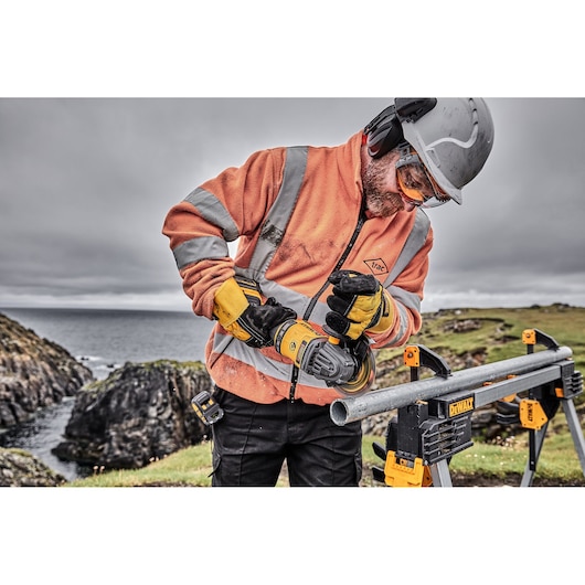 A worker wearing a safety helmet, goggles, earmuffs, and orange high-visibility jacket uses a DEWALT cordless angle grinder to cut a metal pipe outdoors. The pipe is secured on a DEWALT work stand, with a rocky coastline visible in the background.