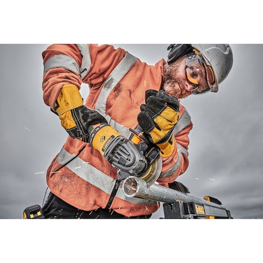A construction worker wearing an orange high-visibility jacket, safety gloves, and a helmet is using a DEWALT cordless angle grinder to cut through a metal pipe, with visible sparks flying.