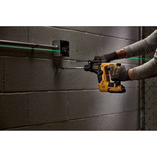 A person wearing gloves uses a DEWALT cordless rotary hammer drill to install or drill into a concrete block wall. The drill is yellow and black, and there is a green laser line visible across metal conduit for alignment.