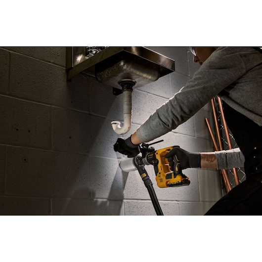 A person wearing gloves uses a DEWALT yellow cordless rotary hammer drill under a sink to work on pipes attached to a concrete wall. The drill is model DCH072L2 and is being used in a plumbing setting.