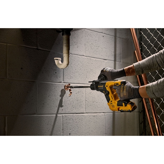 A person using a DEWALT cordless rotary hammer drill to drill into a concrete wall beneath plumbing pipes. The person is wearing gloves and the tool is yellow and black.