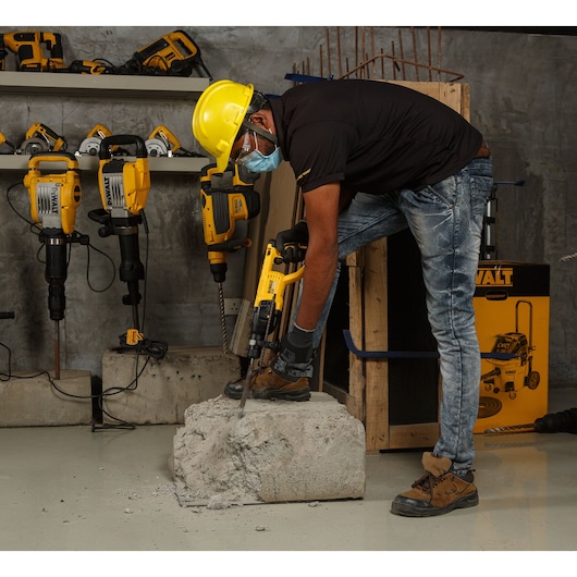Worker uses a DEWALT hammer drill on concrete, wearing safety gear. Other DEWALT tools are visible on shelves.