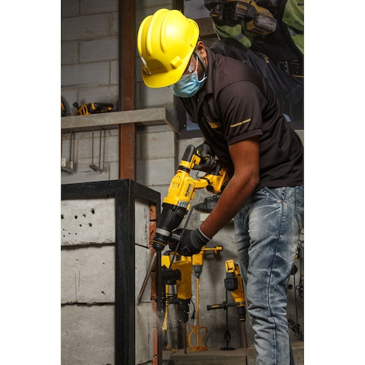 A worker wearing a yellow hard hat, protective glasses, and a face mask is using a DEWALT DCH263P2 power tool to drill into a concrete block in an industrial setting. Other DEWALT tools are visible in the background.