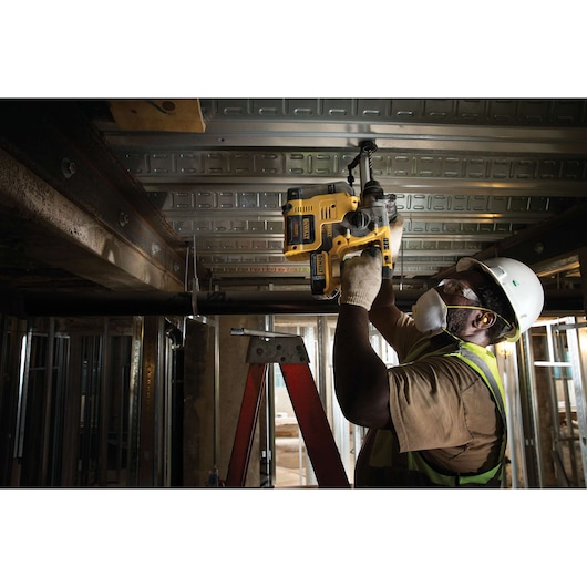 A worker wearing a hard hat, safety vest, gloves, and face mask is using a yellow DEWALT rotary hammer drill (model DCH273P2) to drill into a metal ceiling on a construction site.