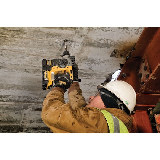 A person wearing a safety helmet, protective glasses, gloves, and high-visibility vest is using a DEWALT cordless rotary hammer drill (model DCH273P2) to drill into a concrete ceiling at a construction site.