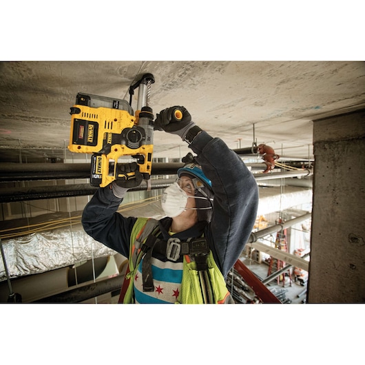 A construction worker wearing safety gear and a face mask uses a yellow DEWALT rotary hammer drill to work on a concrete ceiling at a construction site.