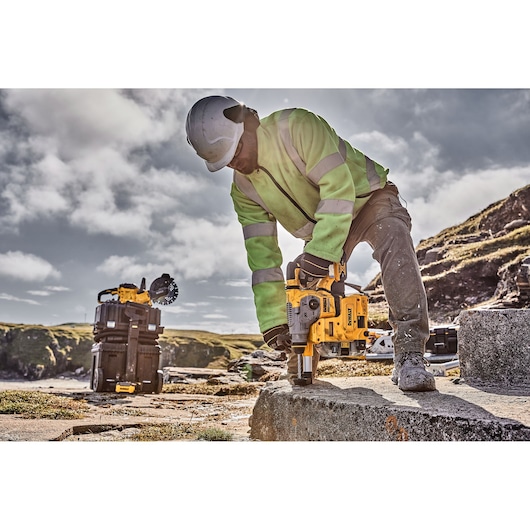 A construction worker wearing a helmet and high-visibility jacket uses a yellow DEWALT power tool to drill into a concrete surface outdoors, with additional DEWALT equipment visible in the background.
