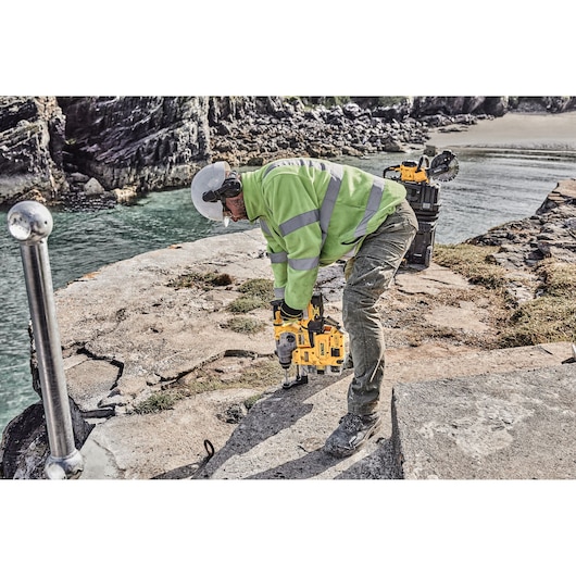 A person wearing a high-visibility jacket, safety helmet, and protective gear is using a yellow DEWALT rotary hammer drill (DCH283P2) on a rocky outdoor construction site near water. A toolbox and additional equipment are nearby.