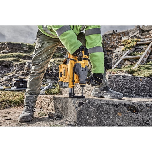 A worker wearing gloves and safety gear is using a yellow DEWALT rotary hammer drill on a concrete slab outdoors. Construction tools and materials are visible in the background.
