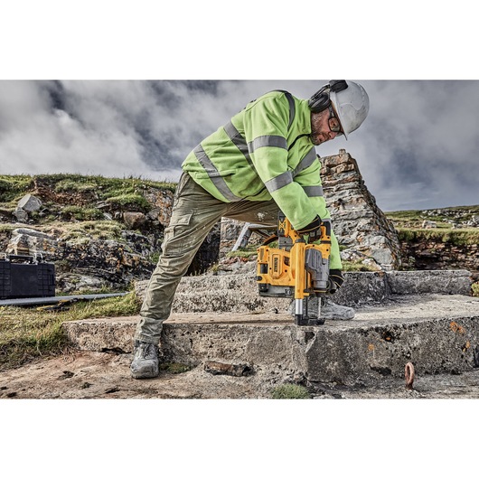 A construction worker wearing safety gear is using a yellow DEWALT rotary hammer drill (DCH283P2) to drill into a concrete surface outdoors. The worker is dressed in a high-visibility green jacket, helmet, gloves, and protective eyewear.