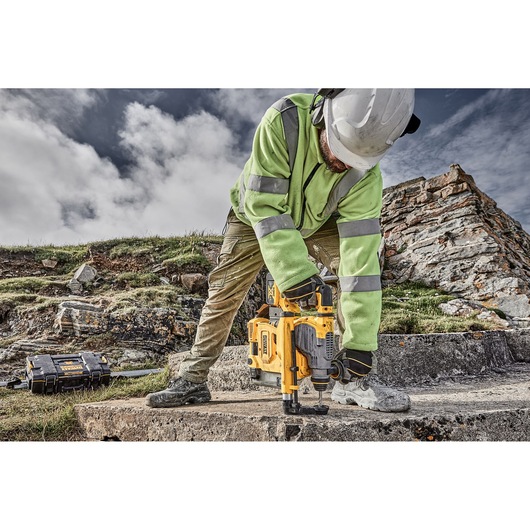 A person wearing a reflective jacket, gloves, and helmet is using a DEWALT yellow power tool on a concrete surface outdoors. There is a rocky landscape and a DEWALT toolbox in the background.