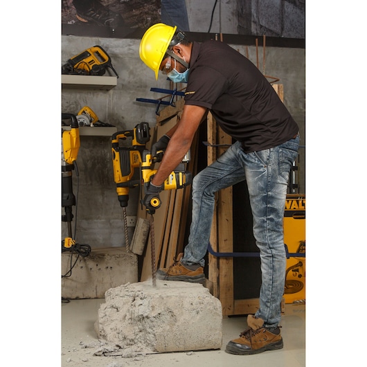 Worker drills concrete block with DEWALT DCH333NT rotary hammer. Wearing safety gear in hardware workshop.