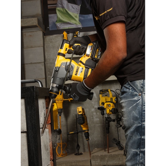 Person using DEWALT DCH333NT rotary hammer drill on a brick wall. Other DEWALT tools visible in the background.