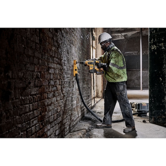 A construction worker wearing a safety helmet and reflective jacket is operating a DEWALT branded power tool to drill into a brick wall in an industrial setting. A vacuum hose is attached to the tool to collect dust.