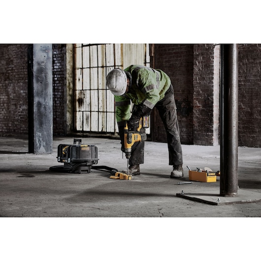A construction worker wearing a helmet and safety jacket is using a DEWALT power tool to drill into a concrete floor in an industrial building. Nearby are a tool case and DEWALT accessories.