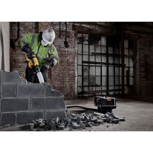 A construction worker in safety gear uses a DEWALT power tool to break a concrete block wall, with rubble scattered on the ground and a DEWALT equipment box nearby in an industrial setting.