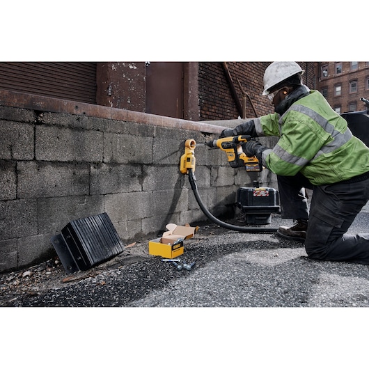 A construction worker wearing a green reflective jacket and white helmet uses a DEWALT cordless rotary hammer drill, model DCH417X2, to drill into a concrete wall outdoors. A toolbox and DEWALT accessories are visible on the ground.