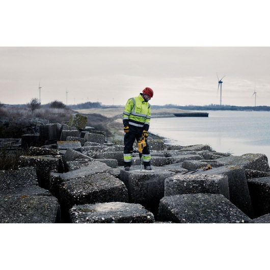 A construction worker wearing high-visibility clothing and a red helmet is using a DEWALT power tool to work on large concrete blocks near a body of water. In the background, wind turbines are visible on a cloudy day.