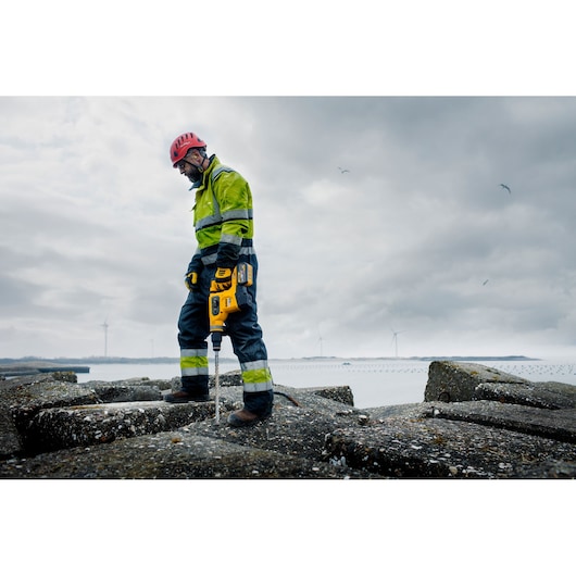 A person wearing a high-visibility jacket, safety helmet, and gloves uses a yellow DEWALT power tool on a rocky surface outdoors. Wind turbines and water are visible in the background under a cloudy sky.