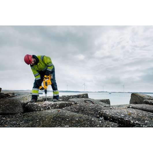 A construction worker wearing a red helmet and high-visibility clothing uses a yellow DEWALT rotary hammer drill (SKU: DCH481N) to drill into large concrete blocks outdoors. Wind turbines are visible in the background under a cloudy sky.