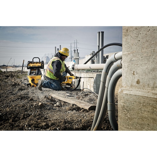 A construction worker wearing a yellow hard hat and safety vest uses a DEWALT power tool to drill into concrete at an outdoor job site. Various pipes and cables are visible, and another DEWALT machine is positioned nearby.