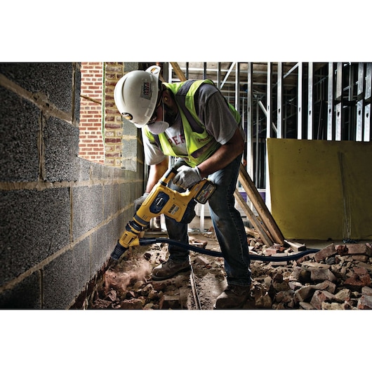 A construction worker wearing a safety helmet and reflective vest operates a yellow DEWALT demolition hammer drill (SKU: DCH481X2) on a brick wall in an industrial building site, with debris and dust visible on the ground.