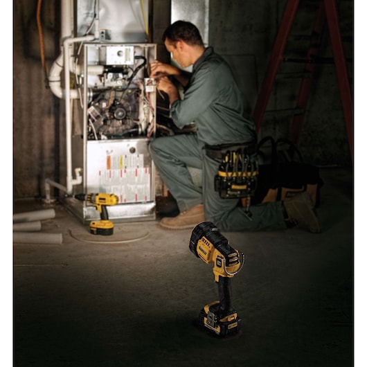 A mechanic is working on a furnace in a dimly lit room. In the foreground is a DEWALT DCL043 cordless spotlight, and next to the furnace is a yellow DEWALT cordless drill. The worker is wearing a green uniform and tan work boots, with DEWALT tools and a tool pouch nearby.