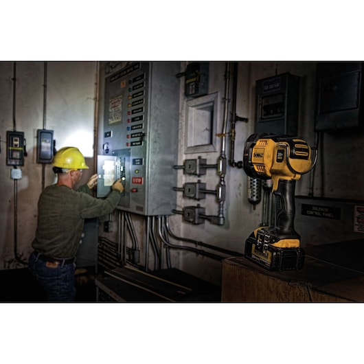 A DEWALT DCL043 LED work light is shown resting on a surface in a dimly lit industrial setting. In the background, a person wearing a yellow hard hat is working with electrical control panels, illuminated by the work light.