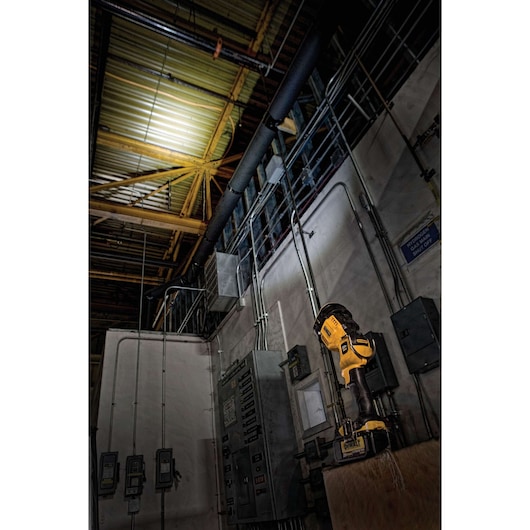 A DEWALT DCL043 handheld LED work light illuminating the ceiling of an industrial electrical room, with various electrical panels and conduit visible.