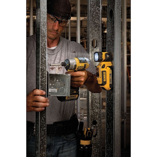 A person using a DEWALT power drill to install a metal electrical box into a steel framing structure. A DEWALT DCL044 portable LED work light is magnetically attached to the metal frame, illuminating the workspace. The person's face is obscured.